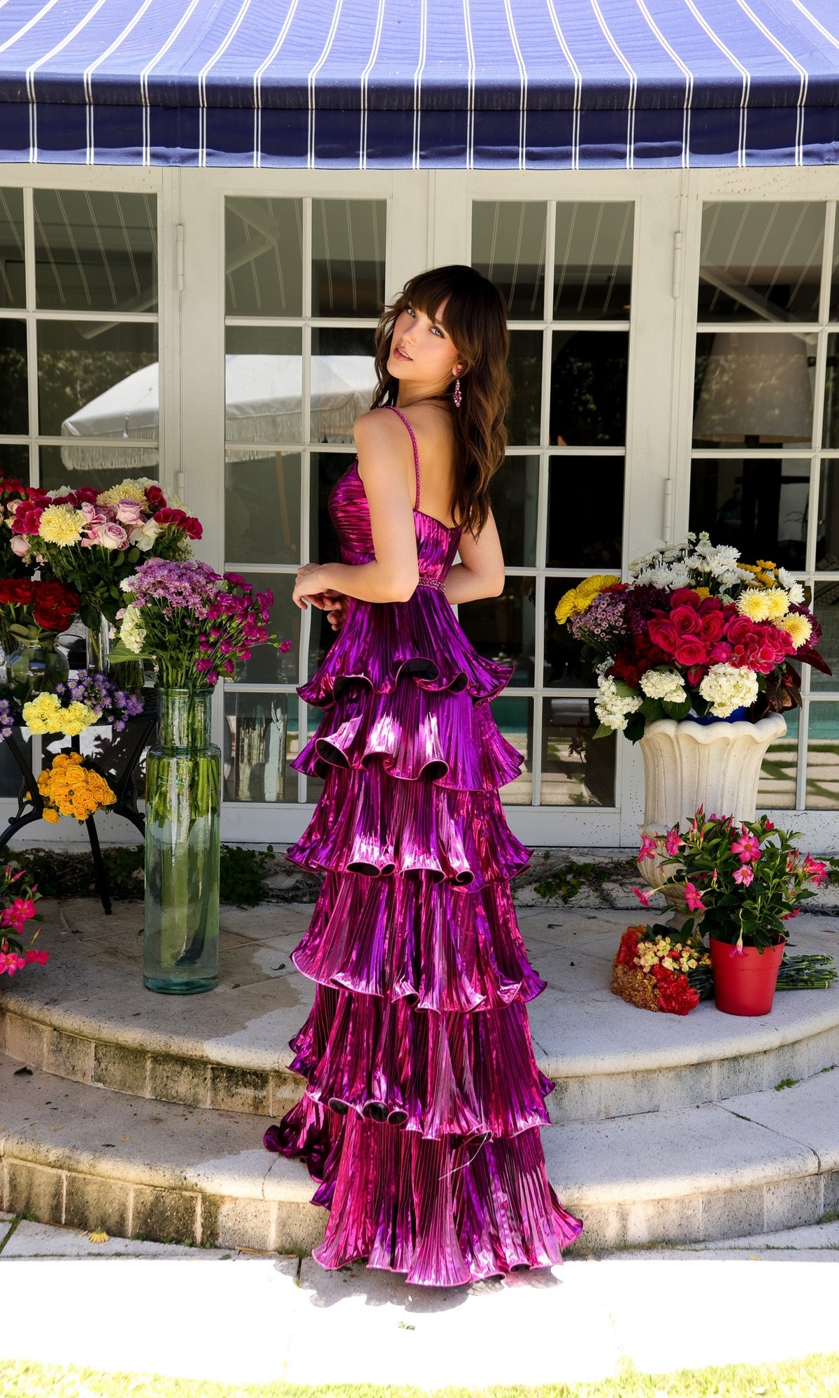 A woman in the Ava Presley 29263 Long Metallic Ruffle Prom Dress stands outdoors by a white building, surrounded by colorful flower arrangements.